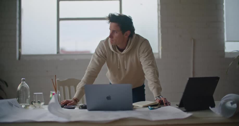 A man stands at his desk in front of his laptop and taps his fingers and he thinks
