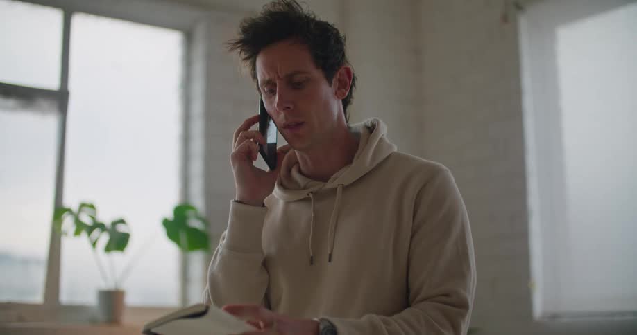 Man standing in room talking on mobile phone consults notebook before leaning down to look at screen