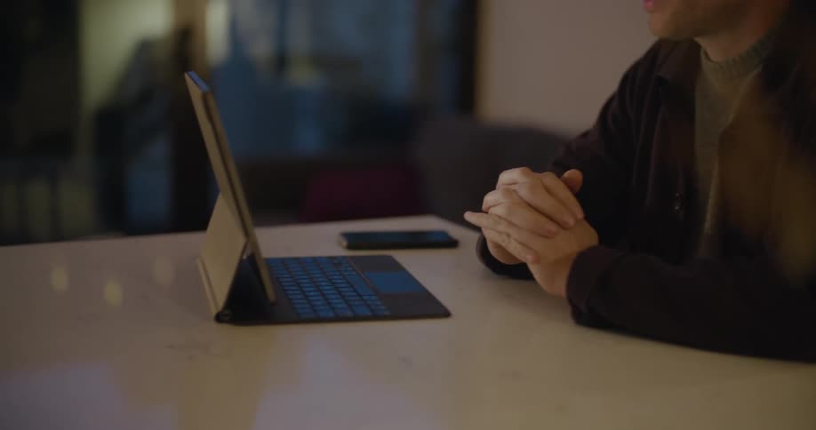A man in a room sitting at a table using a laptop with a wall and window in the background