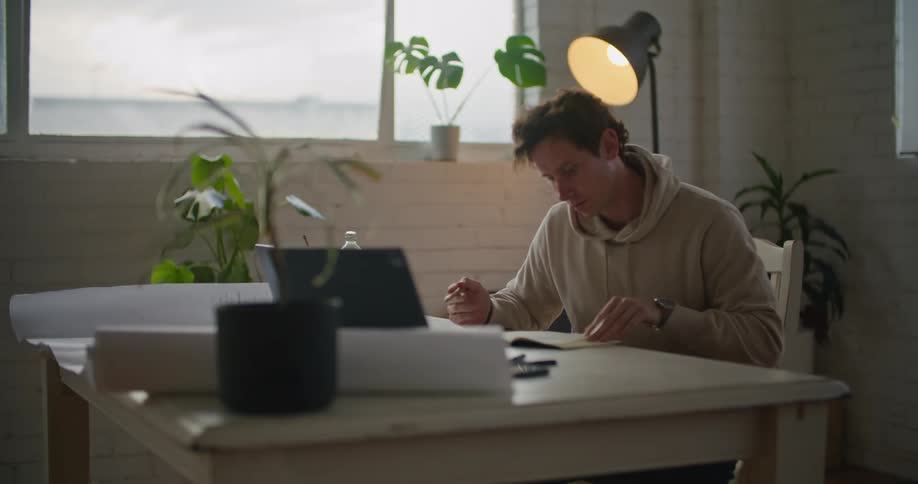 A man sits at a desk with with an electronic device and a book clearly thinking about what hes looking at