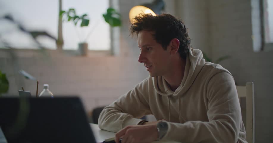 A man in a room sitting at a table using a laptop with a wall and window in the background