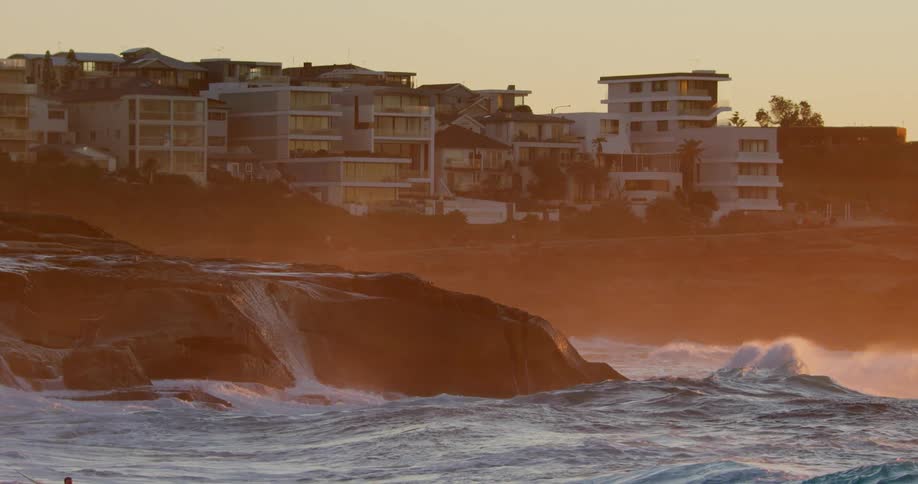 Ocean waves break and splash up on a rocky shore with apartment buildings near by