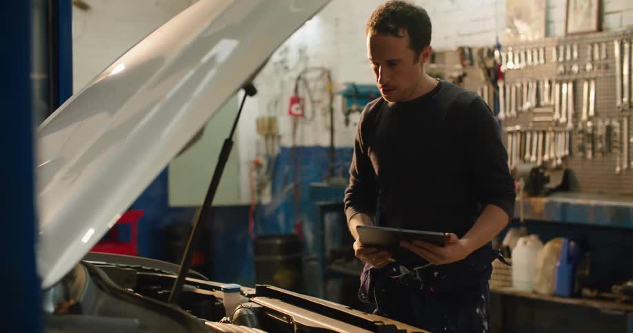 A male mechanic looking under the hood of a car while consulting a tablet
