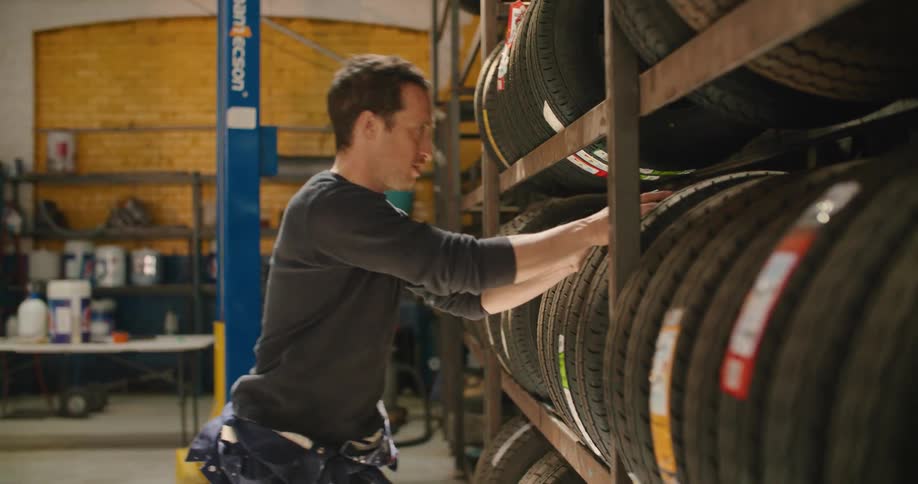 A mechanic in a garage searches a rack then selects a tire pulling it out and rolling it away