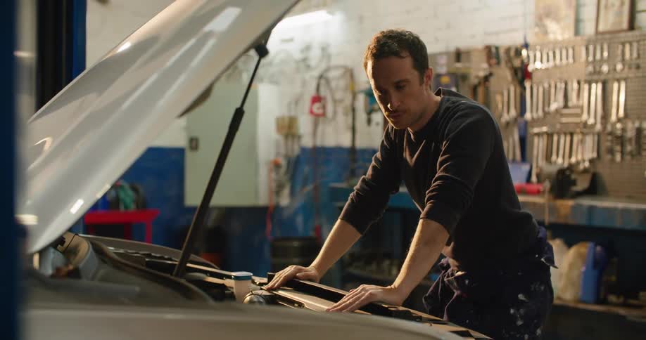 A man in a garage opening the hood of a vehicle with tools and a wall in the background