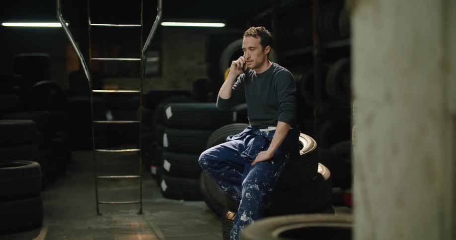 A man in a warehouse sits on a stack of tires and talks on his cell phone