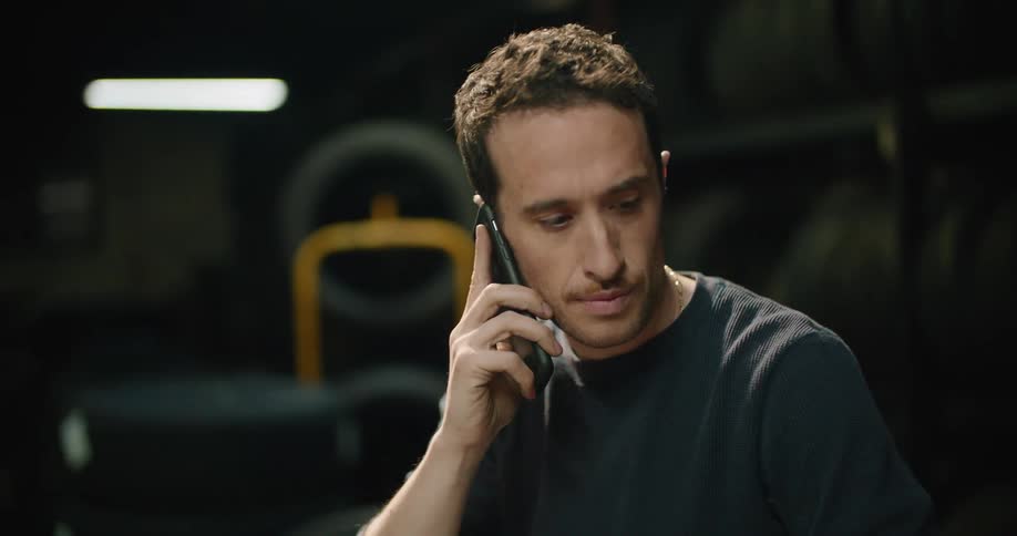 A man in a garage talking on a smart phone with tires and a wall in the background