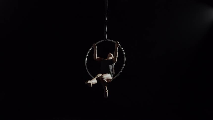A gymnast posing atop a ring hanging from the ceiling does a flip to change position into sitting inside the ring