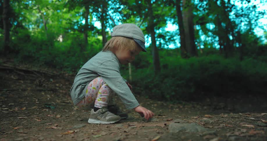 A little girl with a hat touching a rock on the ground with trees the sky and clouds in the background