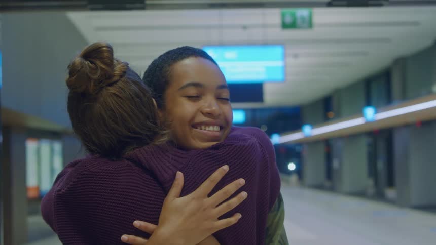 A woman walking through doors in a terminal and hugging another woman with signs in the background