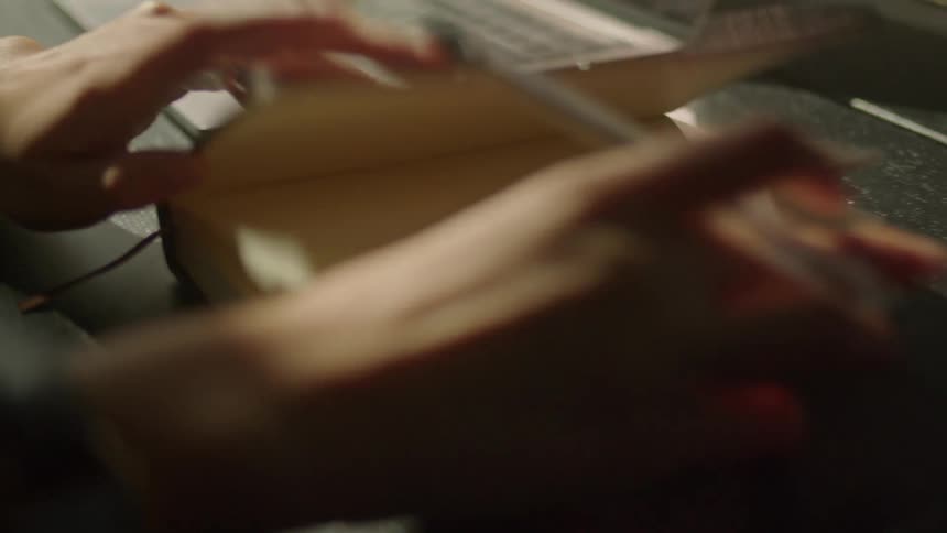 A person in a room taking a pen from a jar and opening a book next to a laptop