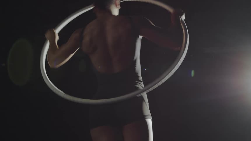 An aerial gymnast pulls herself up onto a spinning hoop twirling as a light shines
