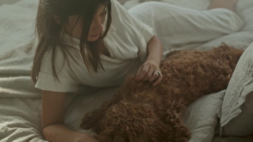 A woman pets a dog while lying next to it in a bed