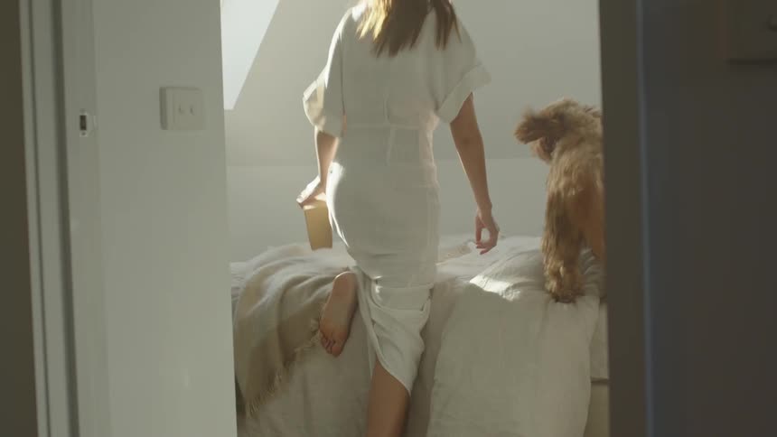 A woman with a book and a dog walk into a bedroom and get on a bed with a window and a wall in the background