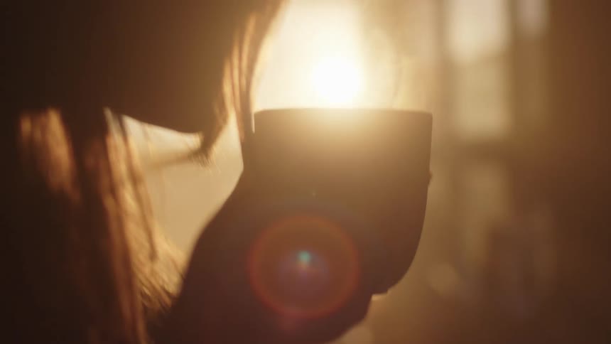 A woman in a room holding a steaming mug with a window and a wall in the background