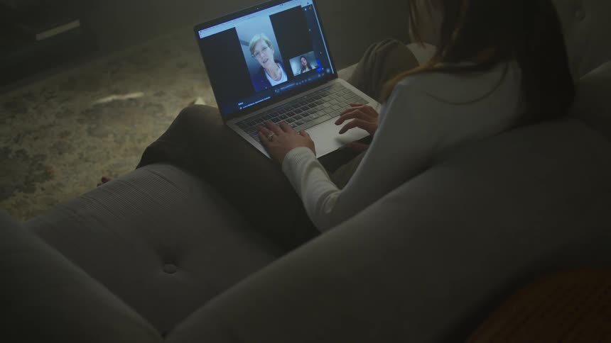 A young woman sits on a couch with her laptop in her lap as she video chats with her mom