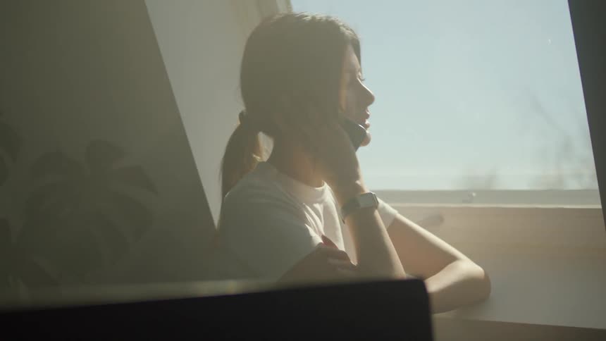A stressed woman takes on her smartphone while standing next to a window in a dimly lit room during the day