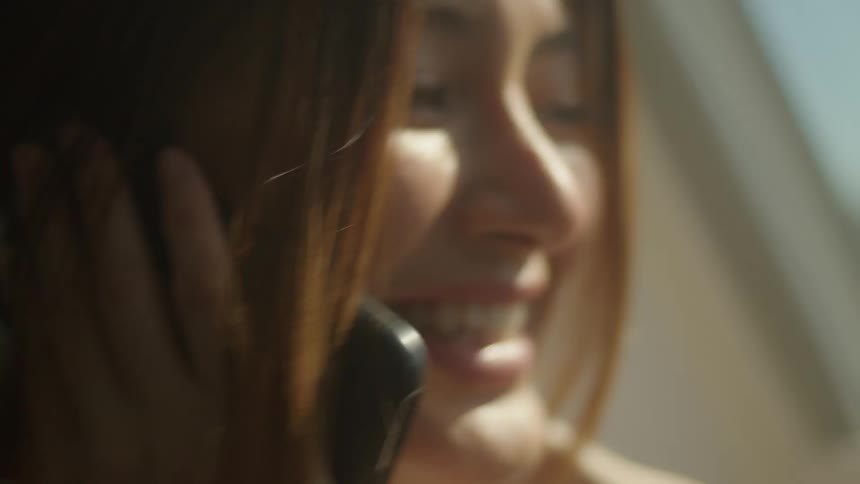 A woman in a room talking on a smart phone with a wall and window in the background