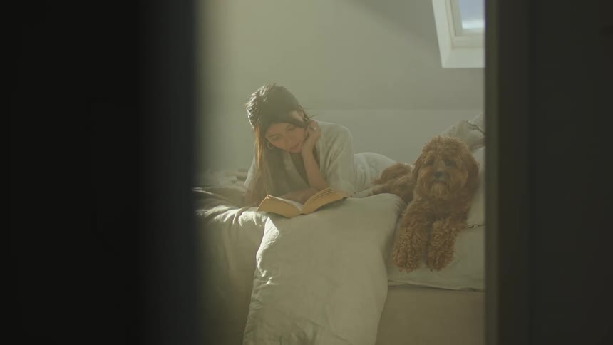 Woman reclines on bed next to furry dog reading