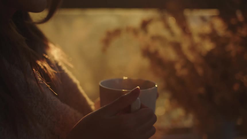 A woman clasps a white glass coffee mug as she stands near a window in low sunlight