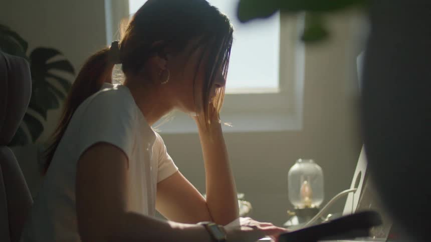 A woman stops looking out a window and types on her laptop