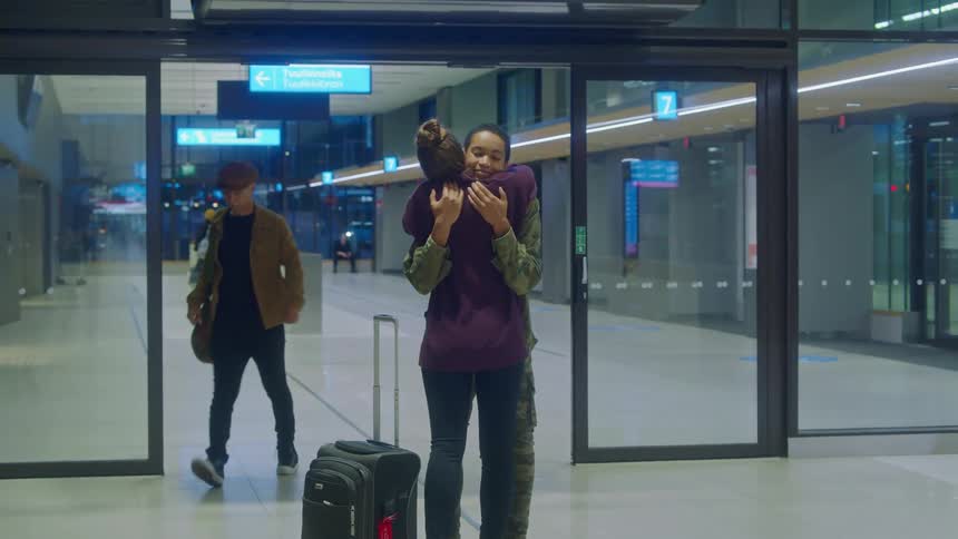 Woman greets arriving woman wearing military fatigues as other travelers move through terminal station automatic doors past them