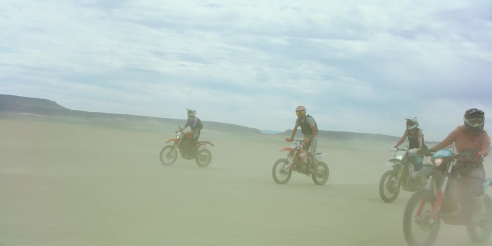 People with helmets riding dirt bikes on a sandy desert with a mountain range sky and clouds in the background