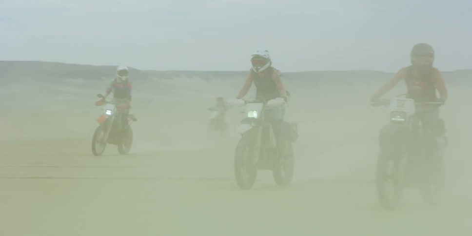 A group of people ride off road motorcycles through a desert in a cloud of dust during the day