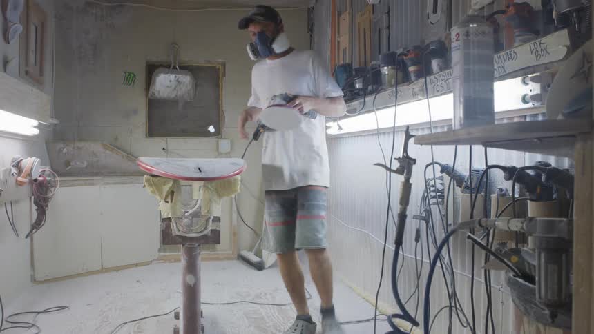 Worker uses a power tool to sand the surface of a surfboard inside a workshop