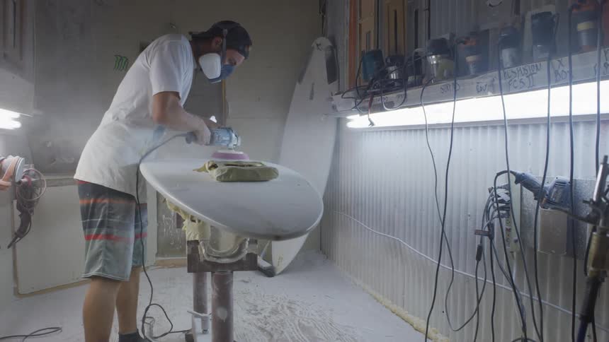 A man in a room with a mask sanding a surfboard with a sander with a wall in the background