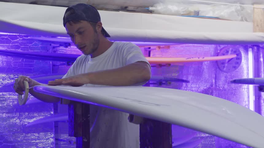 A man with a hat in a room placing tape on a surfboard with a wall in the background