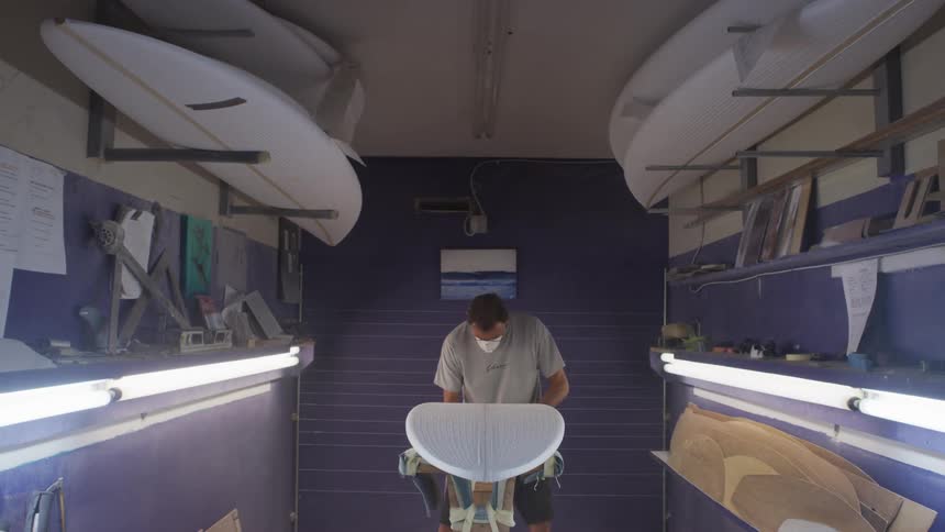 Man in workshop works on surfboard with power tool flipping it over