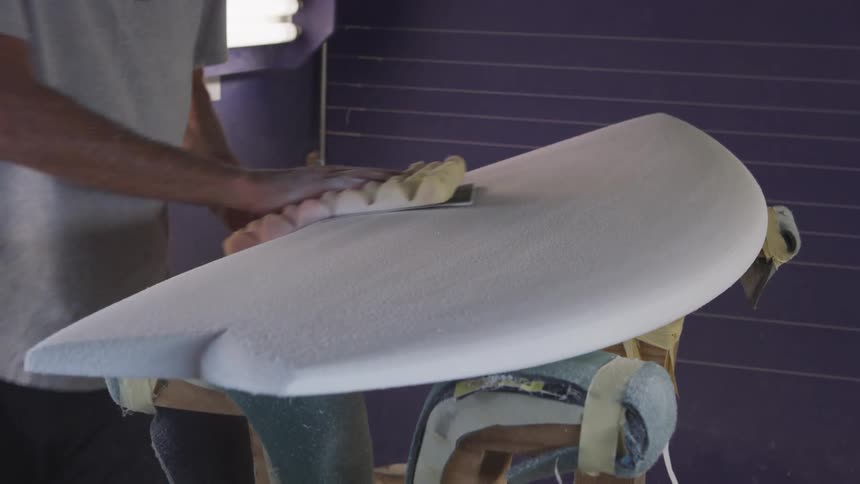 A man in a room sanding a surfboard with sandpaper with a wall in the background