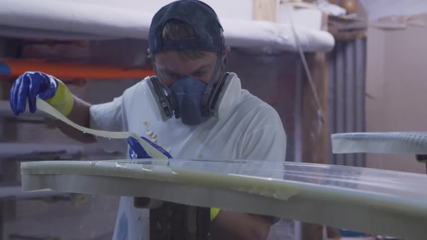 Worker wearing breathing mask and gloves peels tape from the surface of a surfboard inside a workshop
