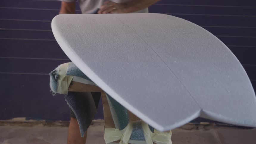A man in a room sanding a surfboard with sandpaper with a wall in the background