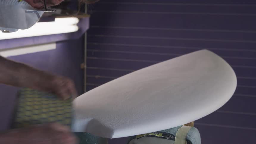 A man uses a large sanding block to smooth the edges of a surfboard carved from wood