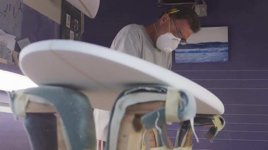 Man dust mask with ventilator sands surfboard in surfboard workshop