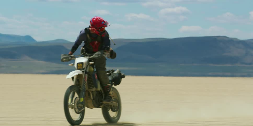 People with helmets riding dirt bikes on a sandy desert with a mountain range sky and clouds in the background