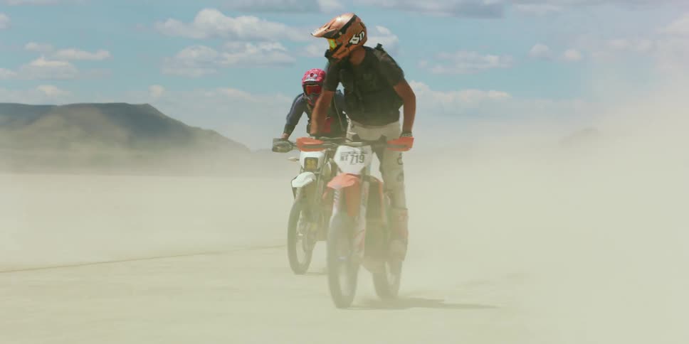 People with helmets riding dirt bikes on a sandy desert with a mountain range sky and clouds in the background