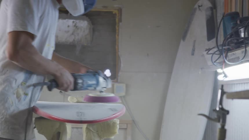 A man in a room with a mask sanding a surfboard with a sander with a wall in the background