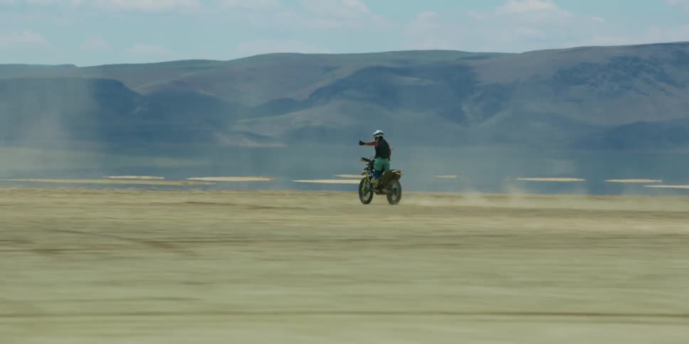 Man riding a motorcycle on a desert kicking clouds of dust near the mountains