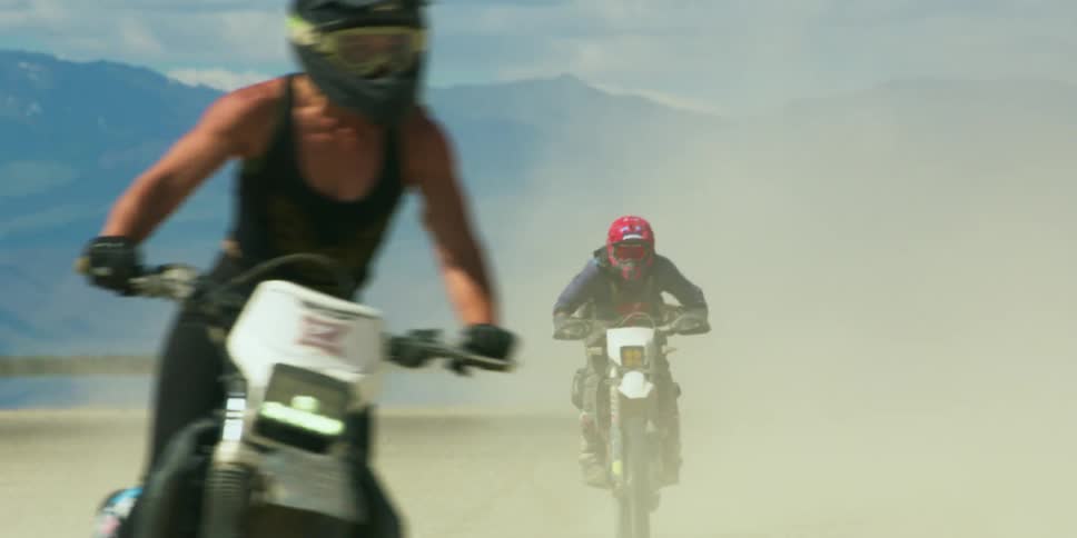 Three cyclists are riding motorcycles on open land with a mountain range behind them
