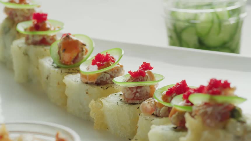 Pieces of sushi on a dish on a table in a room with a jar of sliced cucumbers in the background