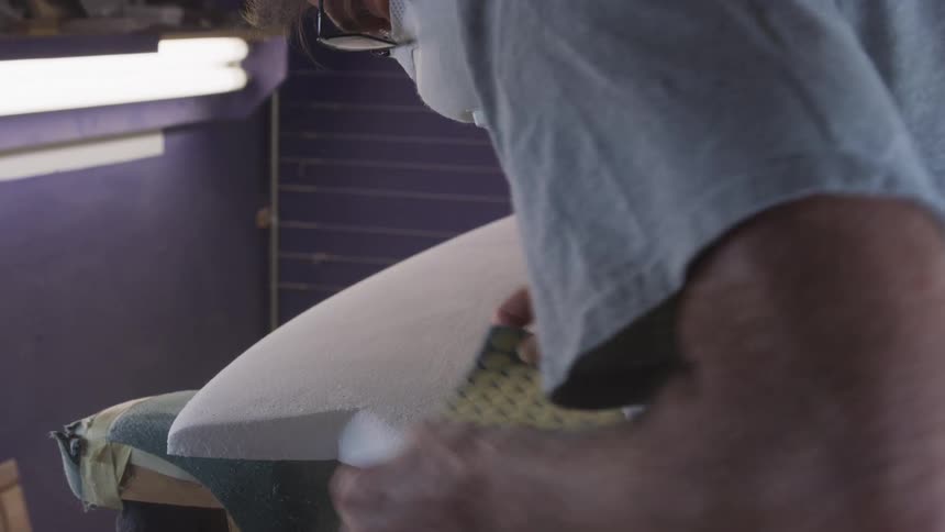 Man sanding the surface of an unfinished surfboard on the workshop