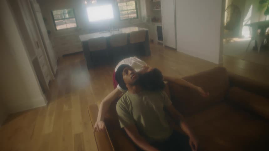 A man sitting on a couch in a room as a woman leans backwoods on him with a wall and a doorway in the background