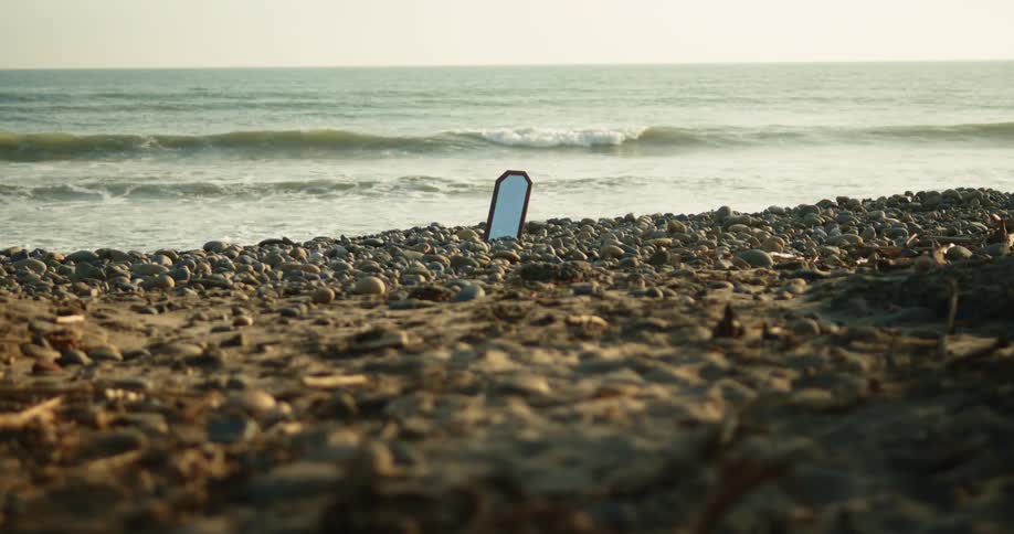 A mirror sits propped up in rocks on the shore as waves roll in