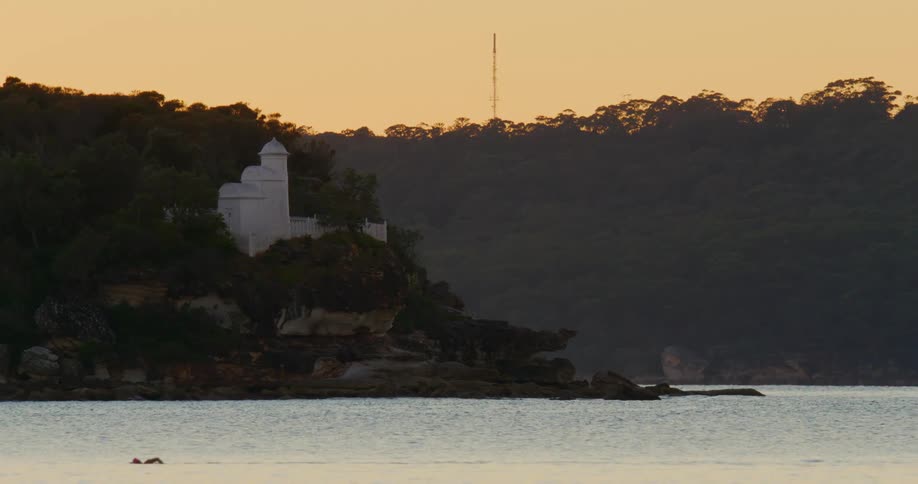 Swimmer swims through ocean passing white building sitting on slope with red sky and forest in background