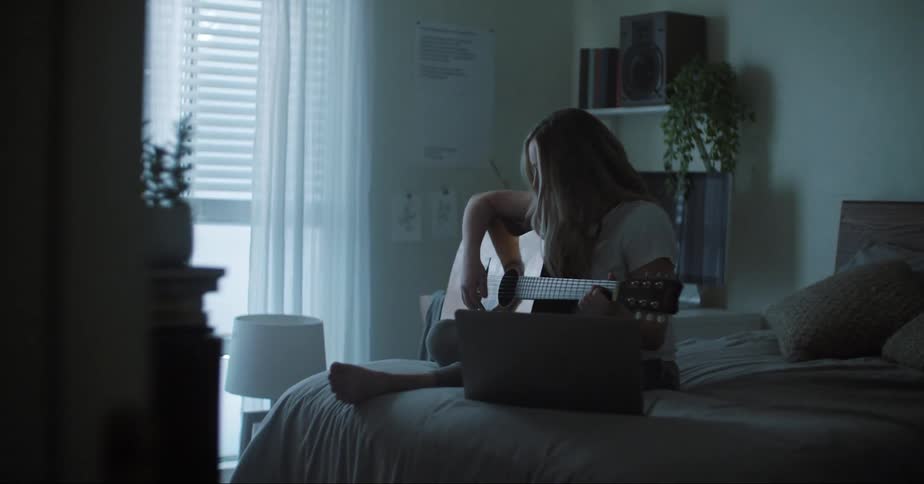 Young blonde girl learning to play acoustic guitar sitting on the bed at home