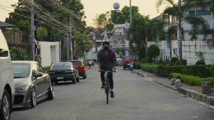 A person bikes down a neighborhood street with cars parked and two people on the side of the road