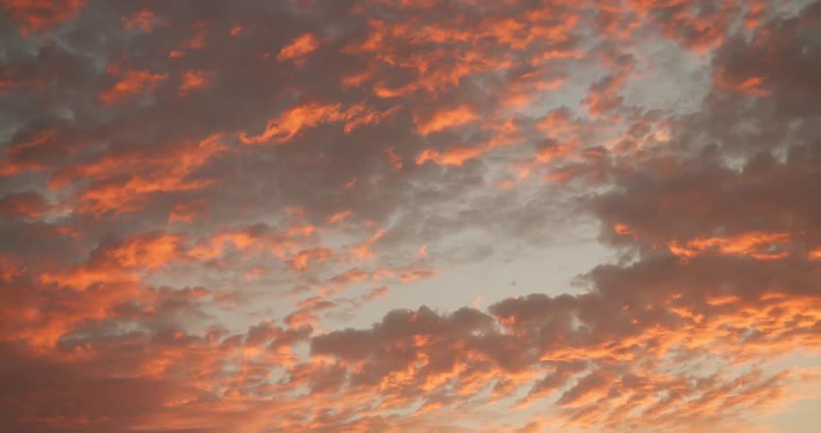 Sun highlights part of clouds turning them crimson while shading other parts dark against a light blue sky